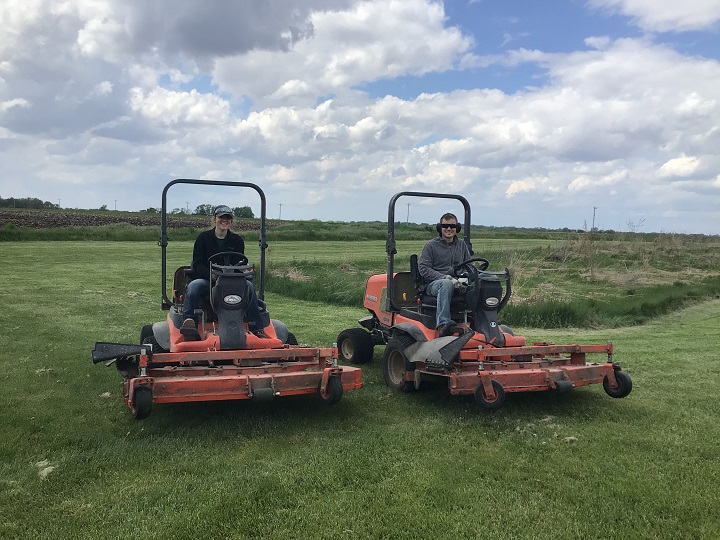 Two summer interns sitting on mowers in a beautiful park with lush green grass and puffy white clouds with little bursts of blue.