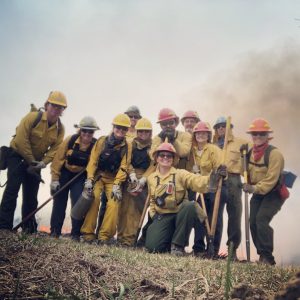 A group of firefighters pose with smoke rising behind.